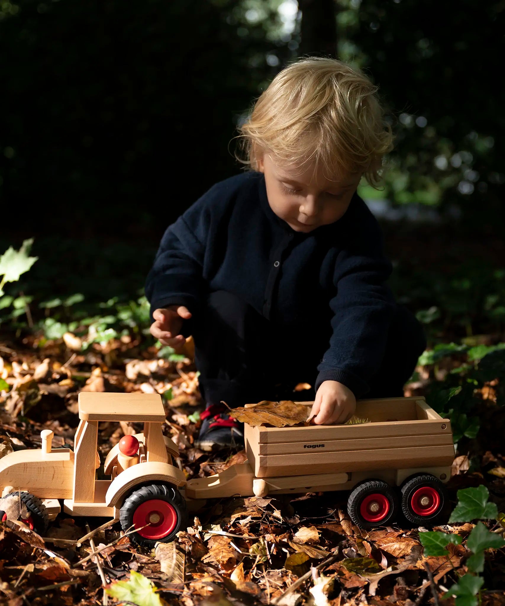 Child playing with the Fagus wooden trailer hitched onto a tractor, filling the trailer with leaves and conkers.