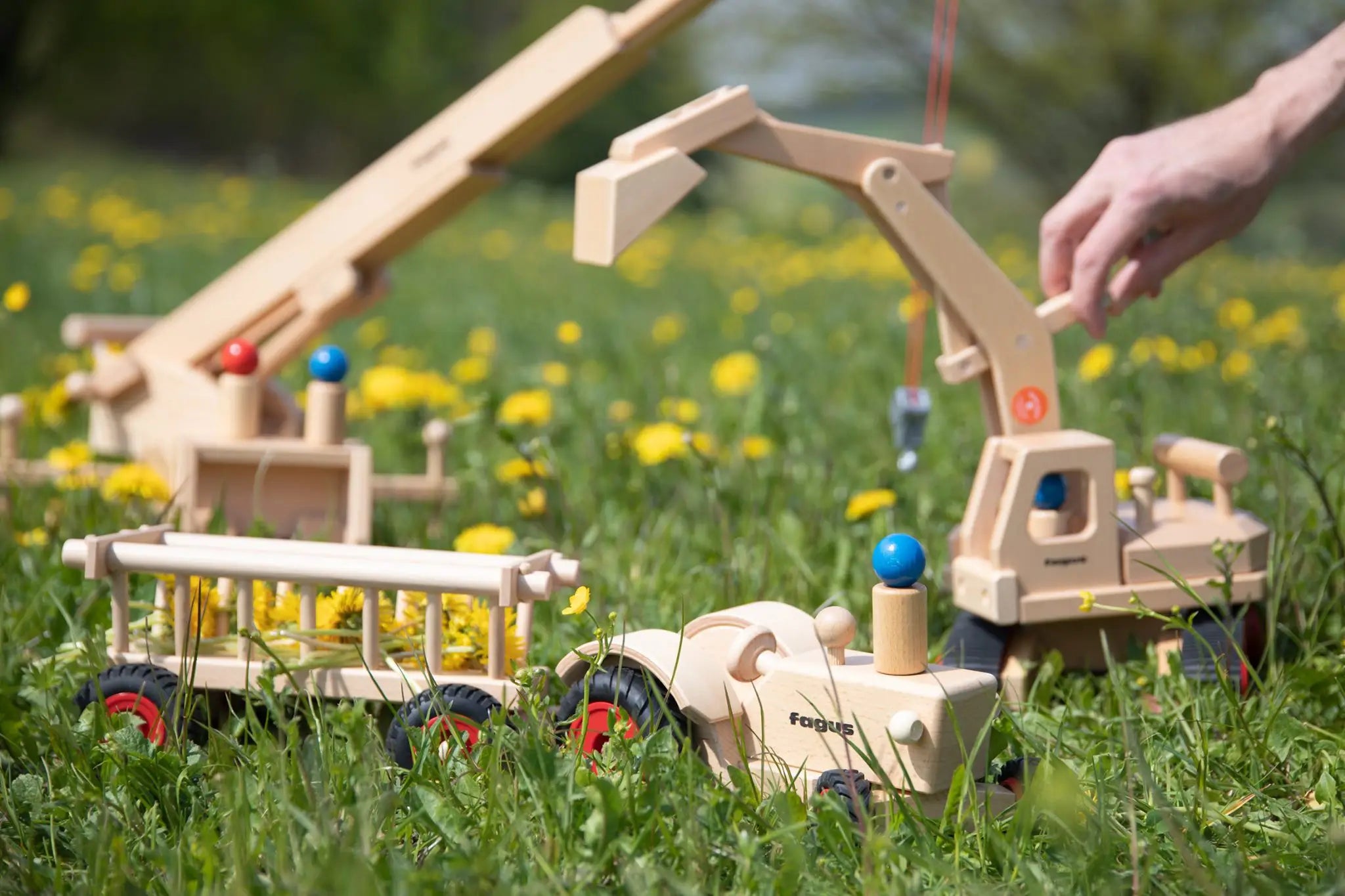 Fagus handmade wooden hay wagon toy attachment hitches onto the tractor with the trailer filled with yellow dandelions. Other Fagus vehicles can be seen in the background.