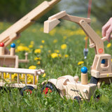 Fagus handmade wooden hay wagon toy attachment hitches onto the tractor with the trailer filled with yellow dandelions. Other Fagus vehicles can be seen in the background.