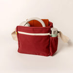 The Fluf organic cotton crossbody lunch bag in vintage burgundy, with a water bottle in the holder, and a pair of headphones inside. 