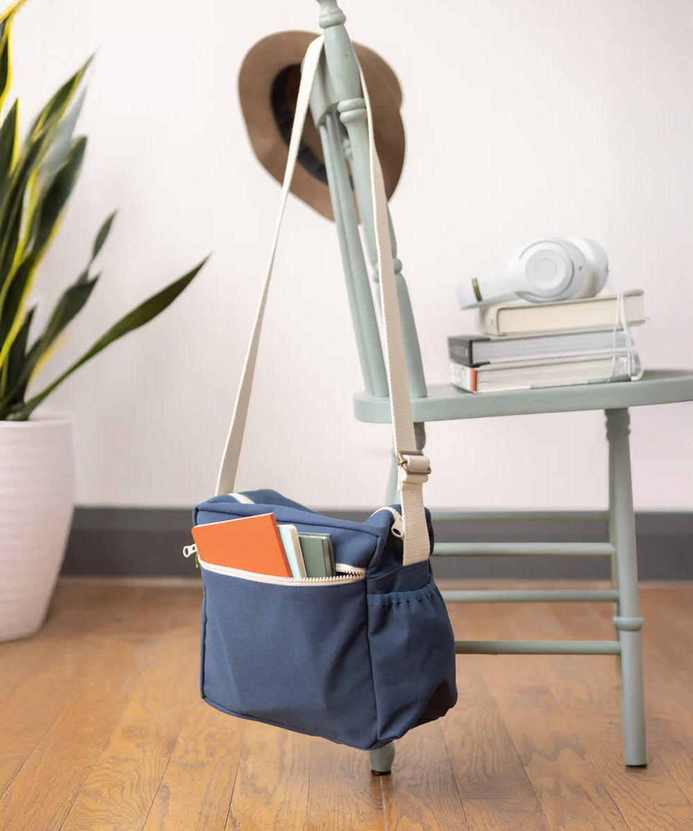 The Fluf organic cotton crossbody lunch bag in navy blue with a white strap, hanging on the back of a chair and filled with books. 