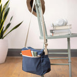 The Fluf organic cotton crossbody lunch bag in navy blue with a white strap, hanging on the back of a chair and filled with books. 