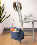 The Fluf organic cotton crossbody lunch bag in navy blue with a white strap, hanging on the back of a chair and filled with books. 