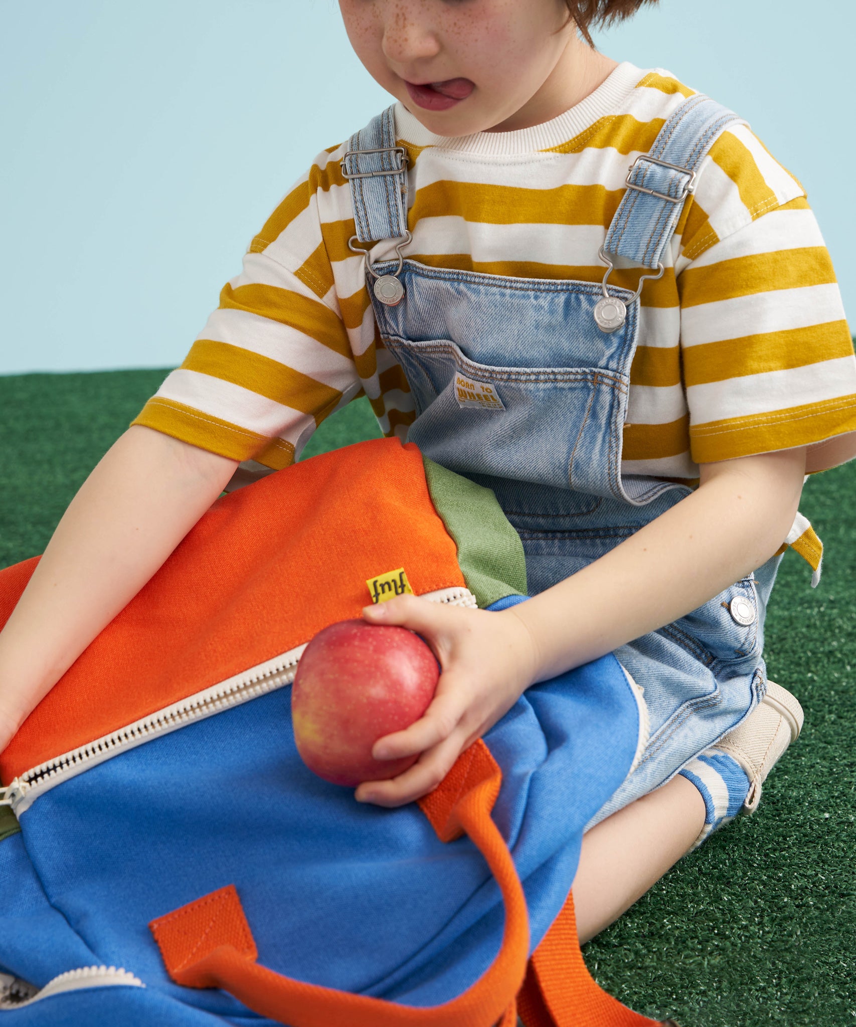 A close up of a child placing an apple in the front zipped pocket on a Fluf kids junior organic cotton backpack. 