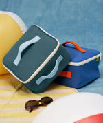 Fluf GOTS organic cotton square lunch bag in a cypress with a white zip detail. Features a light blue carry handle on the top and side. A bag in another colourway has been placed underneath.
