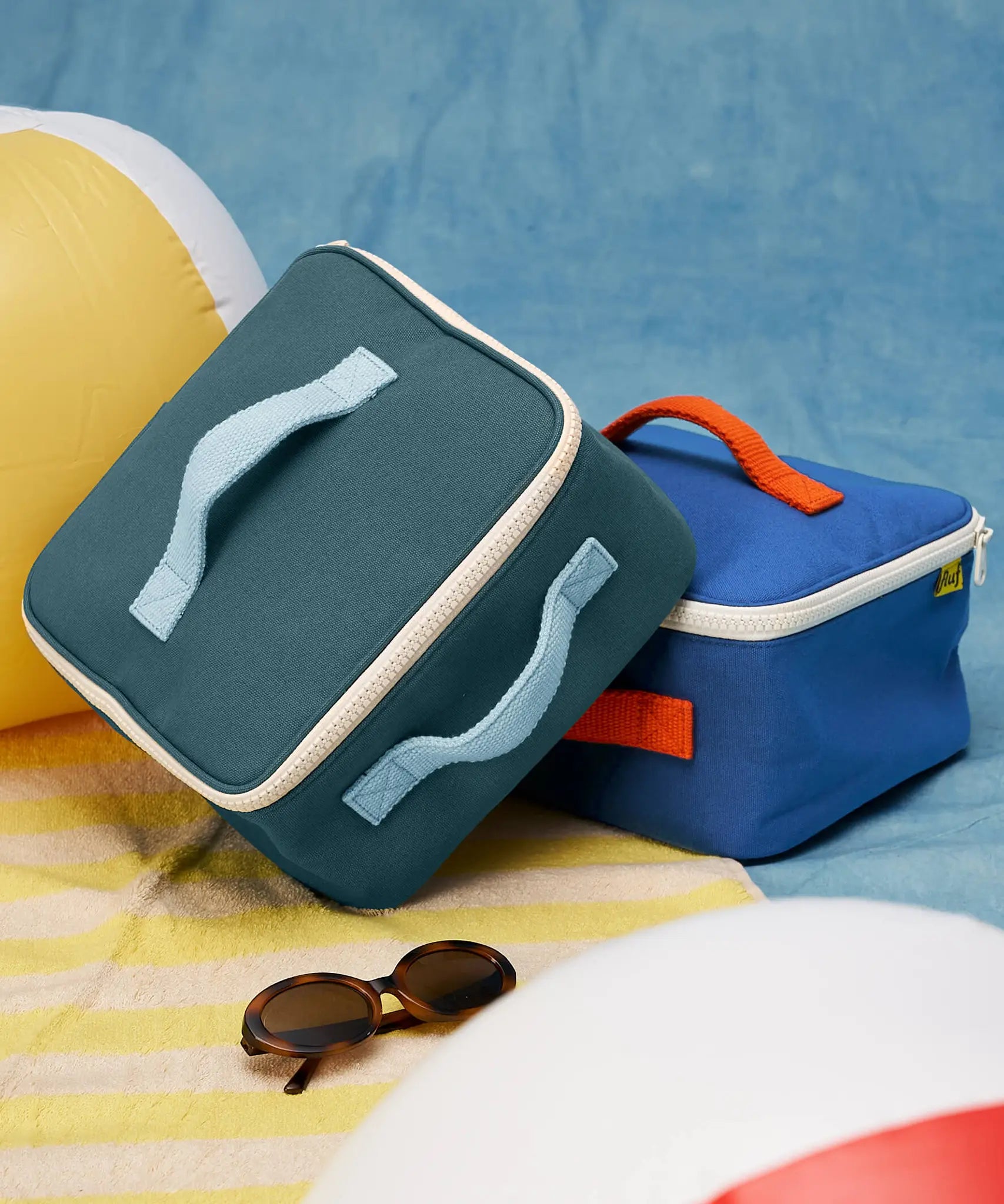 Fluf GOTS organic cotton square lunch bag in a cypress with a white zip detail. Features a light blue carry handle on the top and side. A bag in another colourway has been placed underneath.