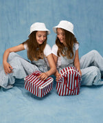 Children sitting, one is holding a Fluf GOTS organic cotton square lunch bag in a awning stripe chili pepper design, the other has a matching zipper lunch bag.