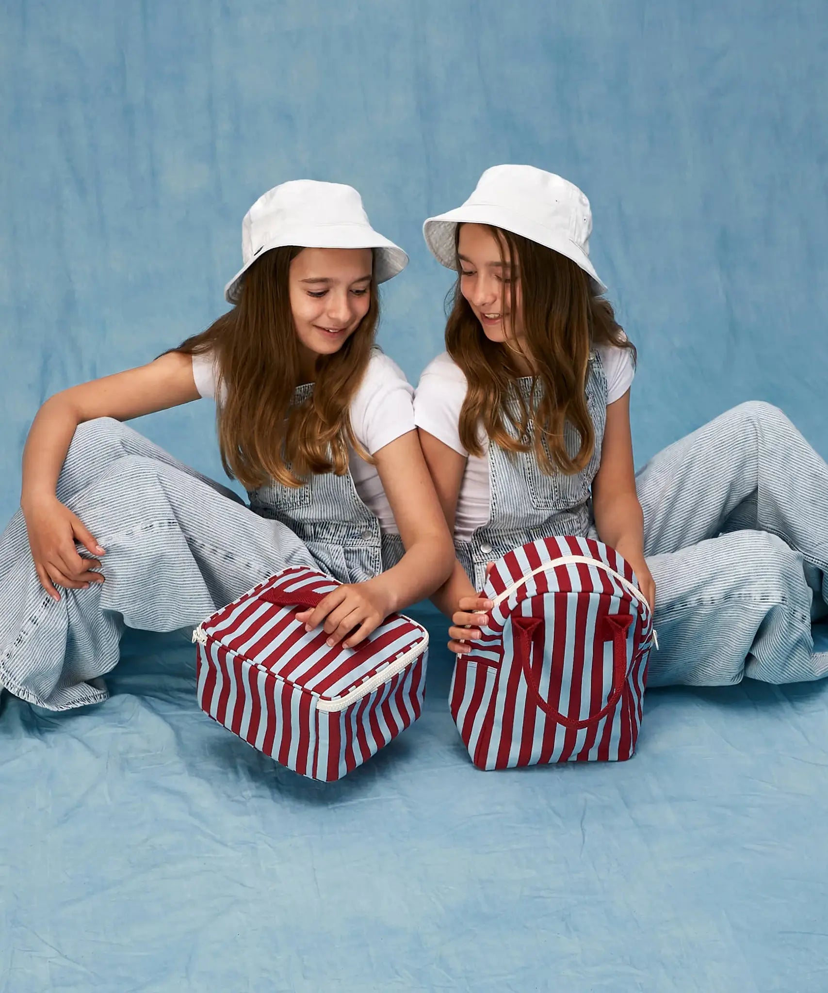 Children sitting, one is holding a Fluf GOTS organic cotton square lunch bag in a awning stripe chili pepper design, the other has a matching zipper lunch bag.