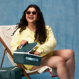 Woman holding a Fluf GOTS organic cotton square lunch bag in a cypress with a white zip detail. Features a light blue carry handle on the top and side.