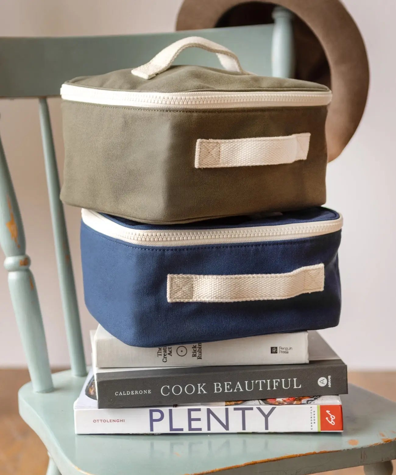 Navy and green Fluf square lunch bags stacked on a chair showing their white zip detail and carry handle on the side