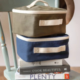 Navy and green Fluf square lunch bags stacked on a chair showing their white zip detail and carry handle on the side
