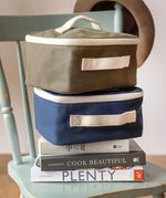 Navy and green Fluf square lunch bags stacked on a chair showing their white zip detail and carry handle on the side