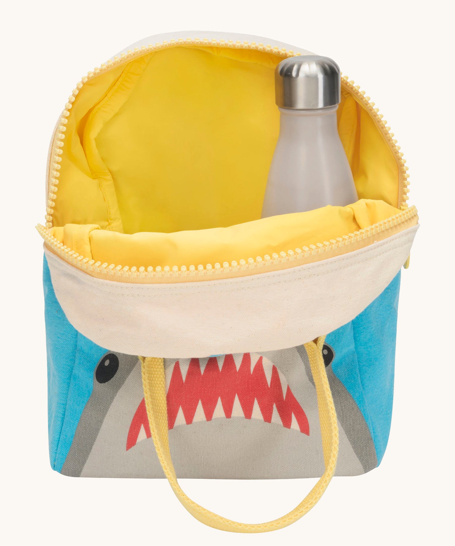 Fluf zip shark lunch bag inside lining in yellow with a bottle packed inside Outside is a blue and grey bag with a design of a shark's head on the bottom with a yellow handle and zip detail on a cream background. 