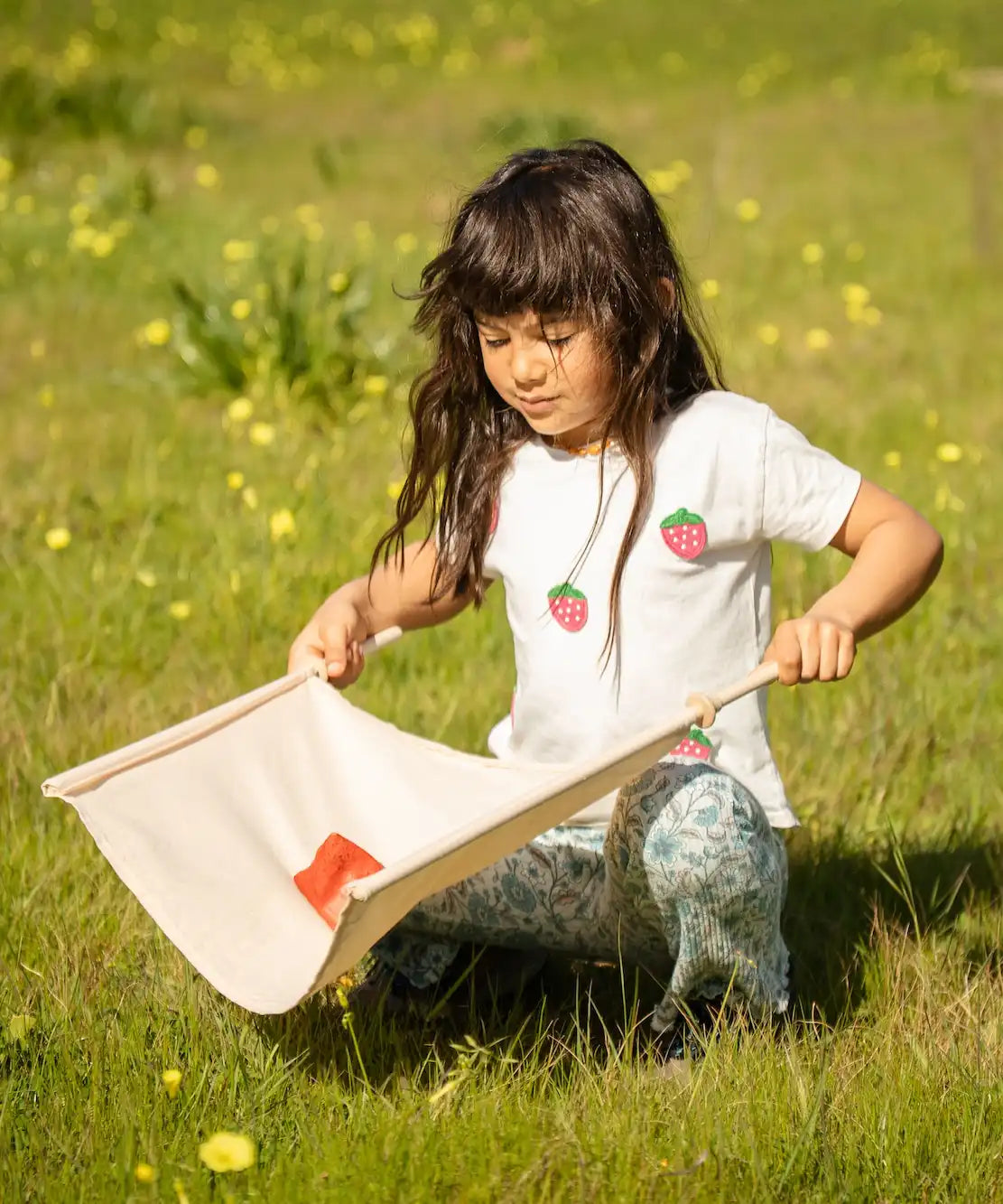 Child playing with the Flup throwing game outdoors, kneeling down holding one of the fabric slings with the throwing bag resting on it. 