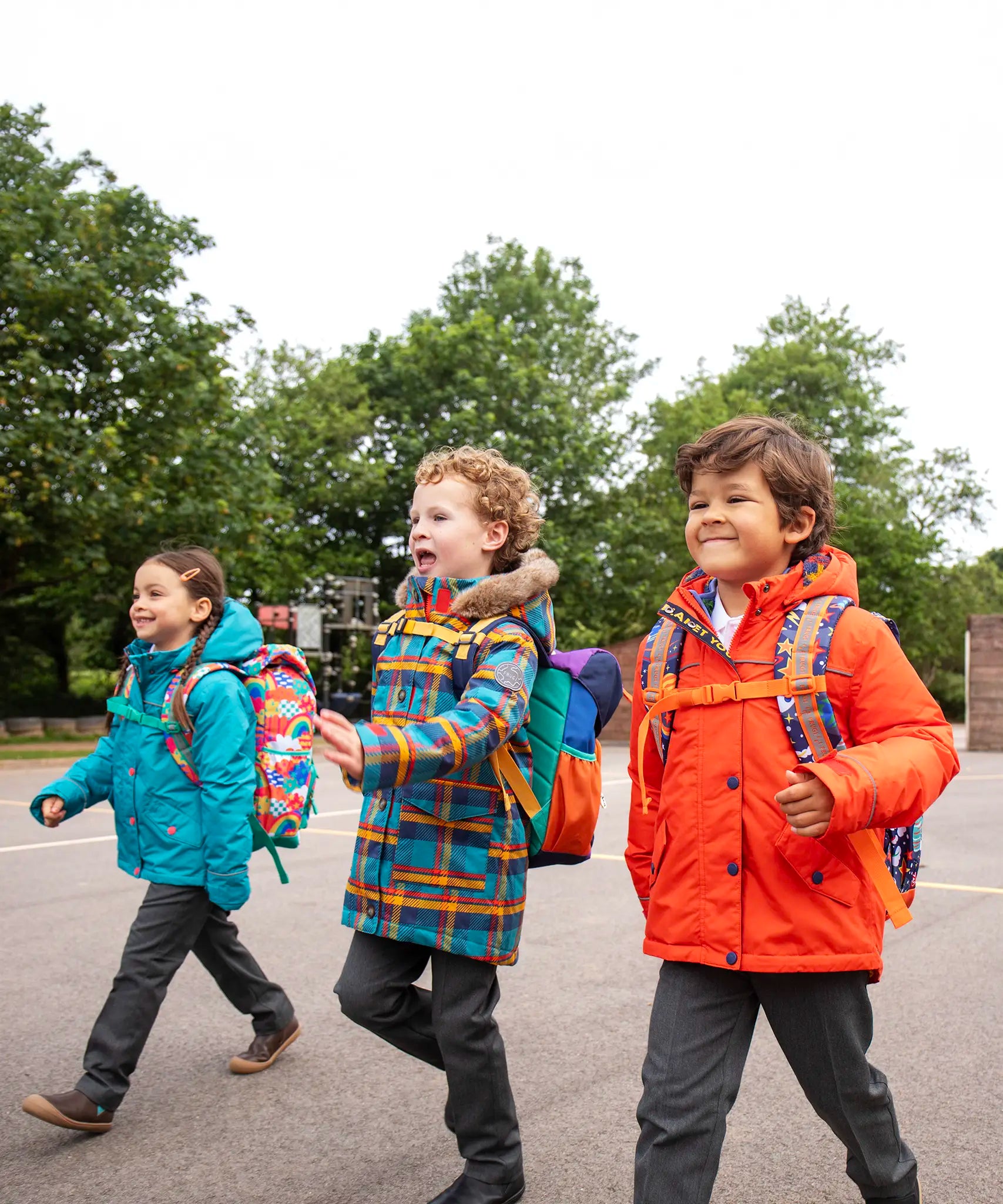 Children walking in a playground all wearing Frugi winter coats. One is wearing the penhale parker, one wearing the fire orange 3-in-one rambler coat and one wearing the boat blue 3-in-one rambler coat.