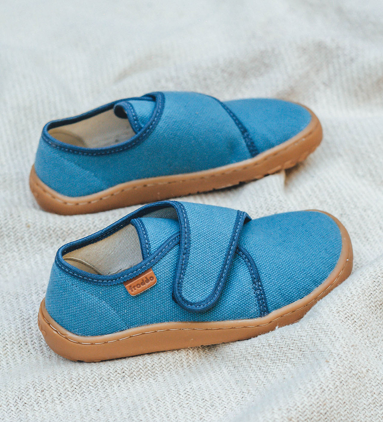 Froddo barefoot kids slipper in denim blue available at Babipur standing on a soft pale blanket. A canvas style slipper shoe in a light neutral colour with a velcro fastener for a secure fit and a dark brown grippy sole