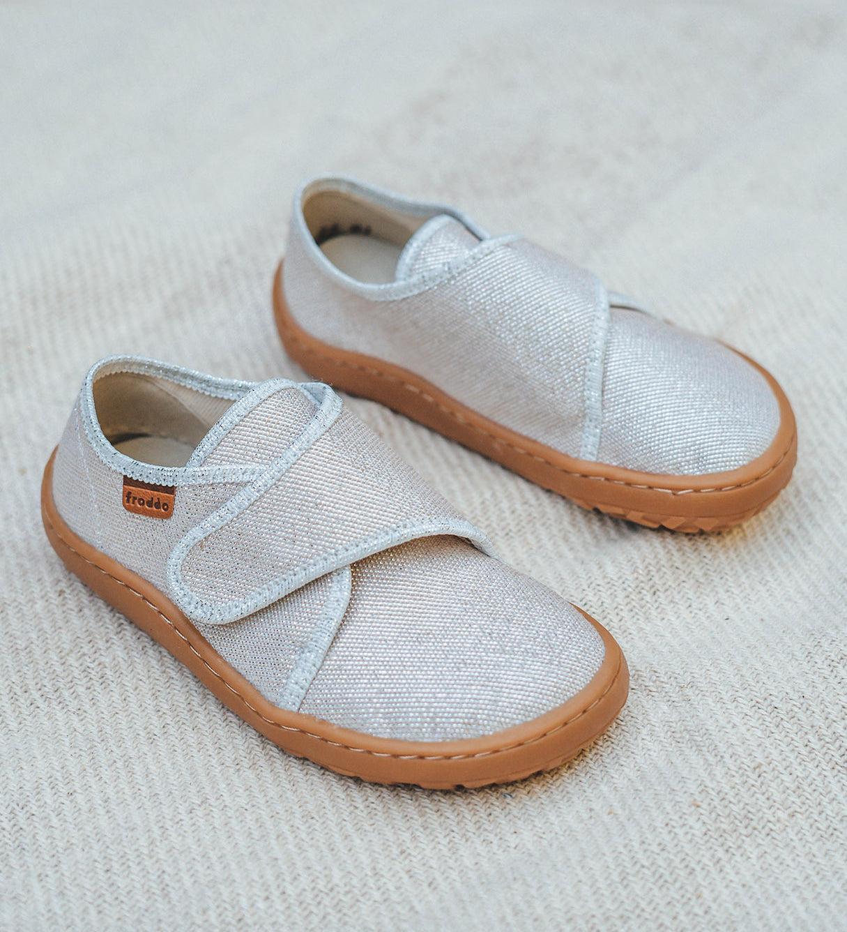 Froddo barefoot kids slipper in gold shine available at Babipur standing on a soft pale blanket. A canvas style slipper shoe in a light neutral colour with a velcro fastener for a secure fit and a dark brown grippy sole