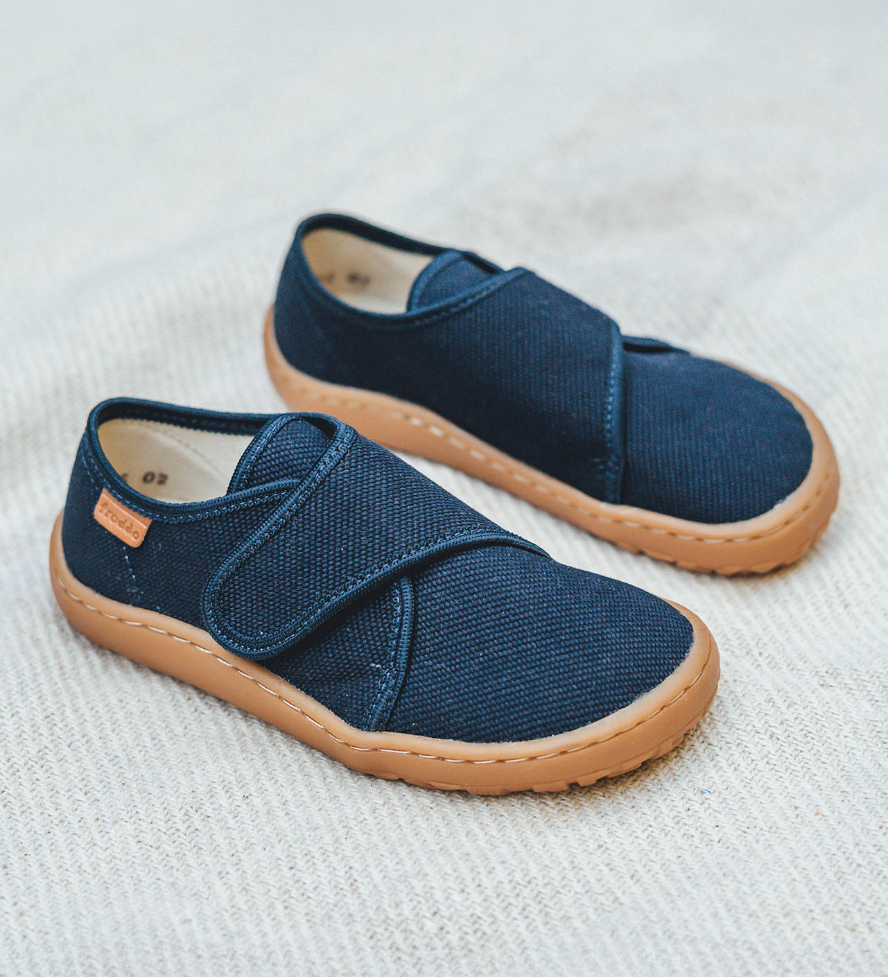 Froddo barefoot kids slipper in dark blue available at Babipur standing on a soft pale blanket. A canvas style slipper shoe in a light neutral colour with a velcro fastener for a secure fit and a dark brown grippy sole