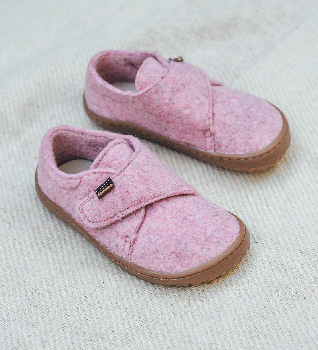 Froddo barefoot wooly children's shoe in pink standing on a pastel coloured blanket. A natural wooly shoe with a velcro fastener for a secure fit and a dark brown grippy and available at Babipur