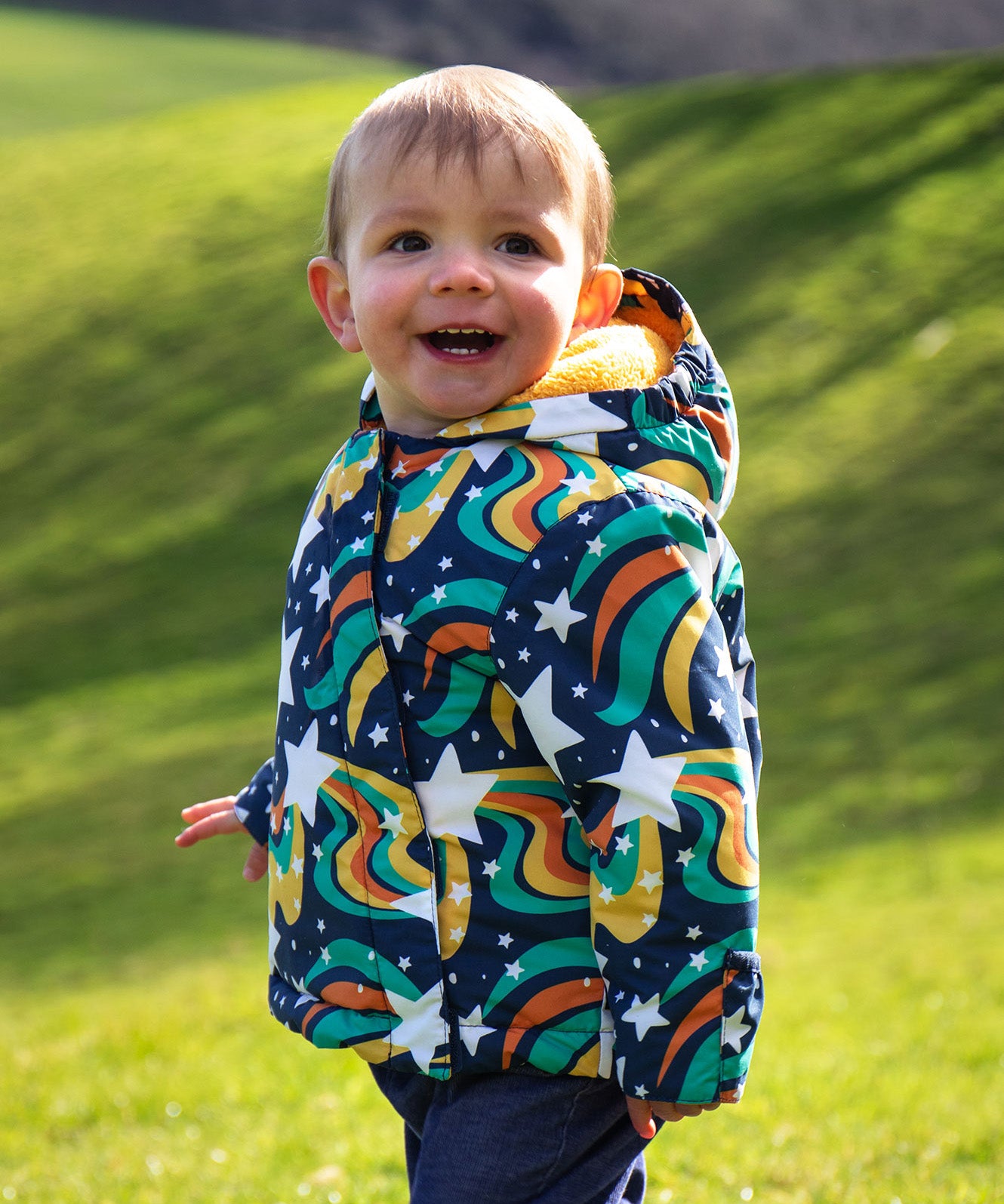 A child wearing the coat from the Frugi 2 in 1 rambler pram suit to coat in magic stars. A thick warm hooded coat with a yellow fleece inner for warmth 