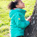 A child wearing the Frugi dragon/mushroom waterproof rambler coat. A thick and warm post consumer recycled polyester coat in bright turquoise with multicoloured popper fasteners and a removable hood