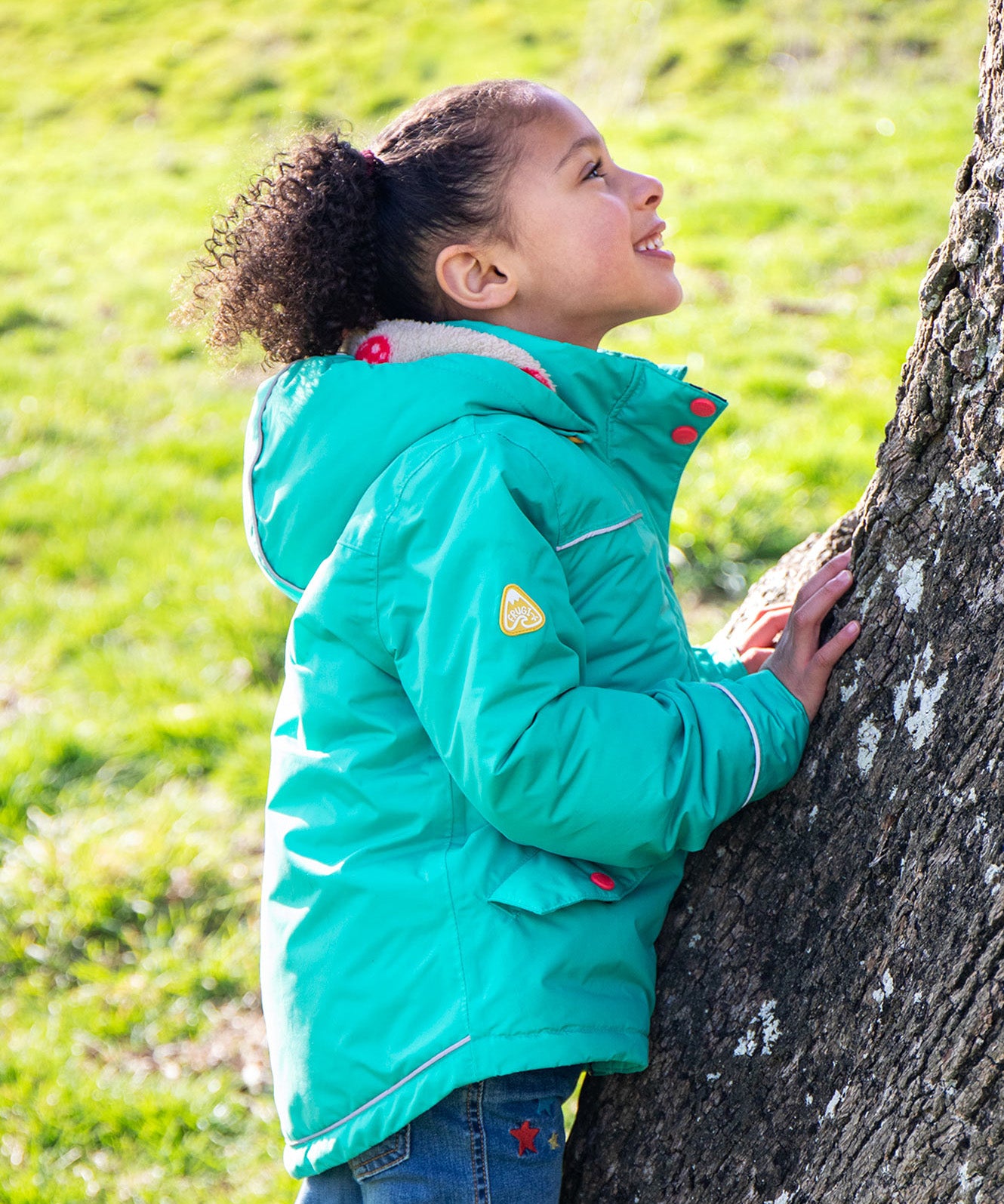A child wearing the Frugi dragon/mushroom waterproof rambler coat. A thick and warm post consumer recycled polyester coat in bright turquoise with multicoloured popper fasteners and a removable hood