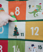 Close up of a green Grapat Nin wooden peg doll being placed into one of the pockets on the Frugi forest festive fun organic cotton advent calendar.