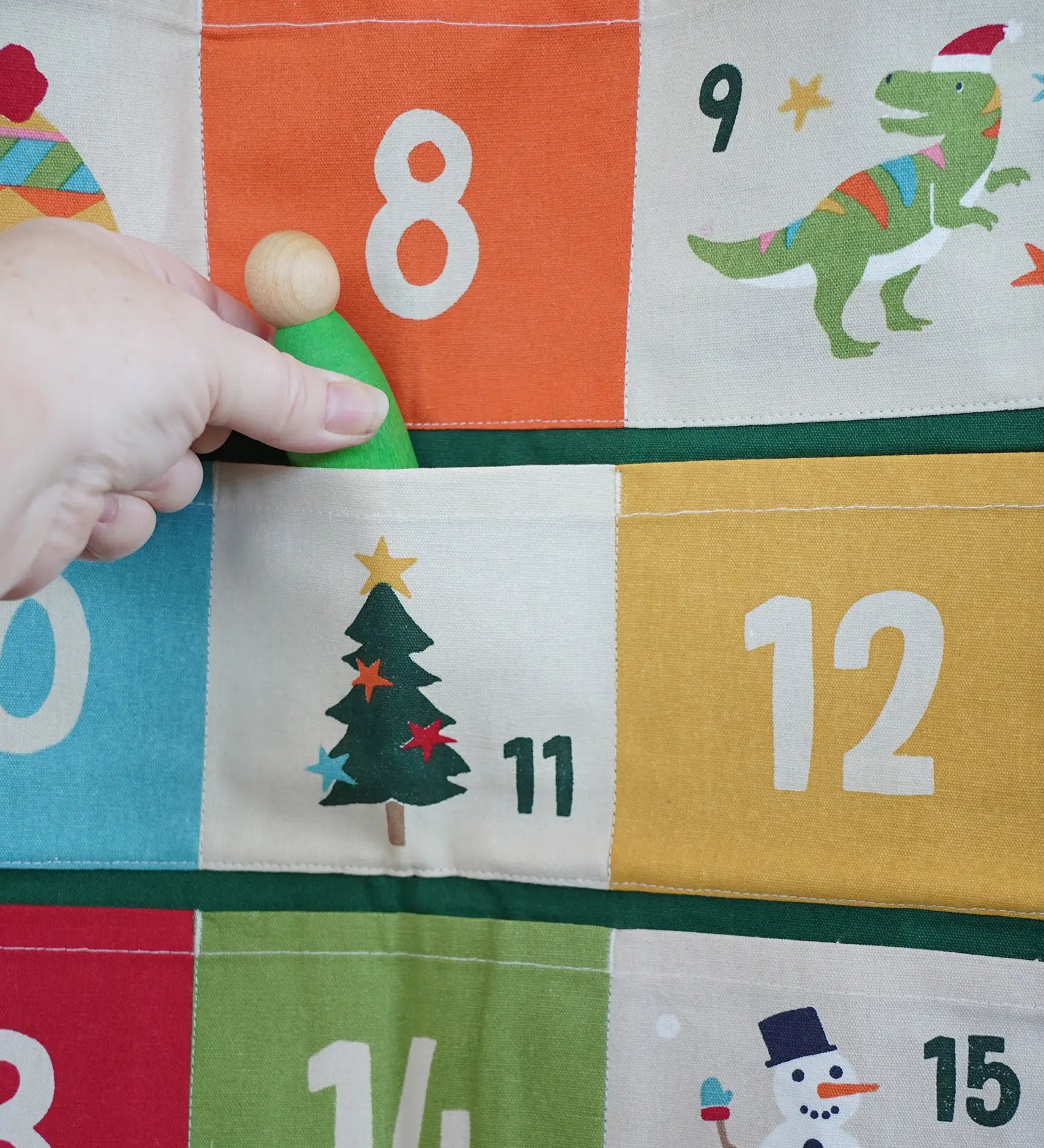 Close up of a green Grapat Nin wooden peg doll being placed into one of the pockets on the Frugi forest festive fun organic cotton advent calendar.