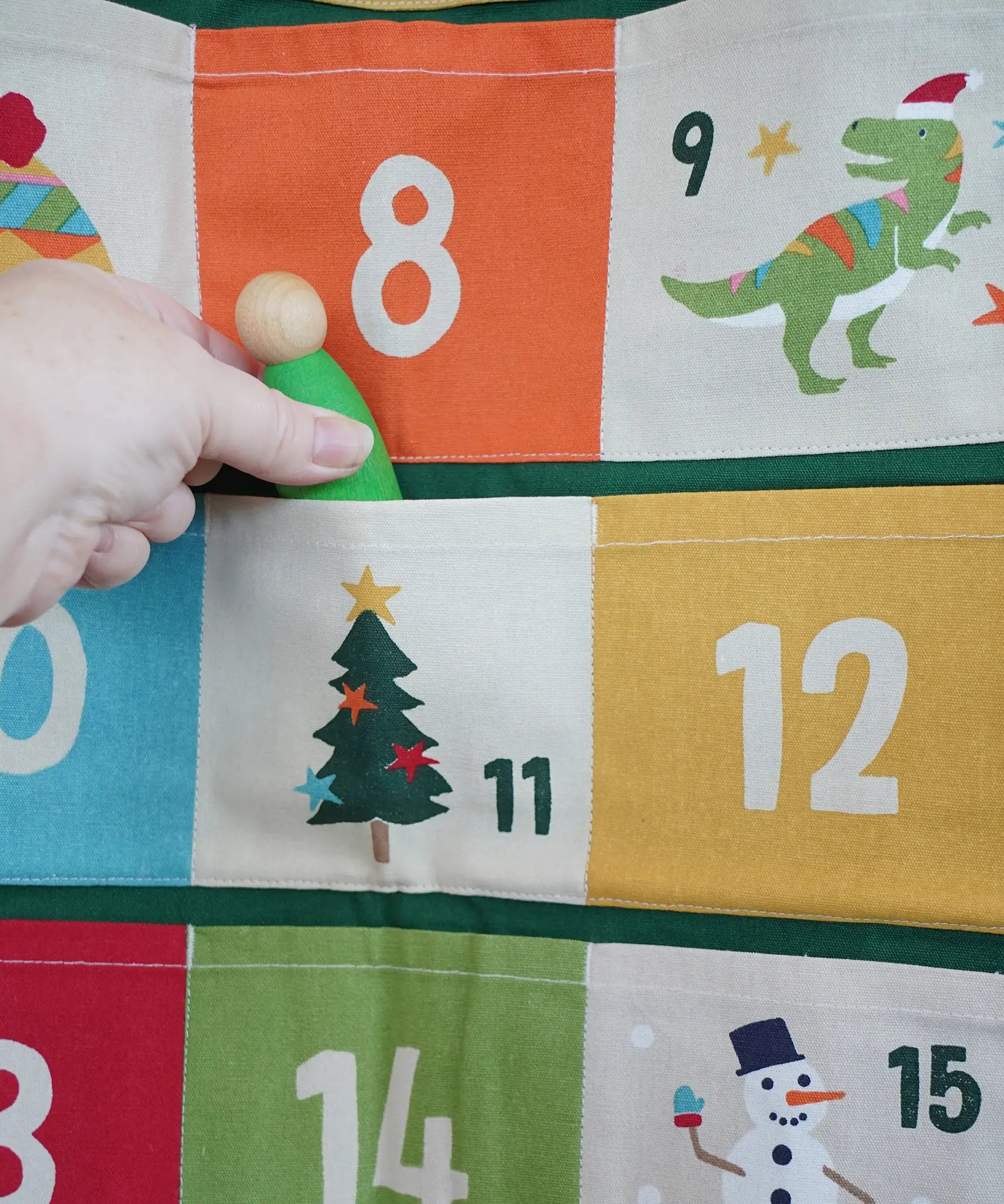 Close up of a green Grapat Nin wooden peg doll being placed into one of the pockets on the Frugi forest festive fun organic cotton advent calendar.