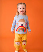 Child wearing the Frugi's GOTS 100% organic cotton top in light blue showing the cute house on a rainbow applique on the front with matching yellow character applique joggers. 