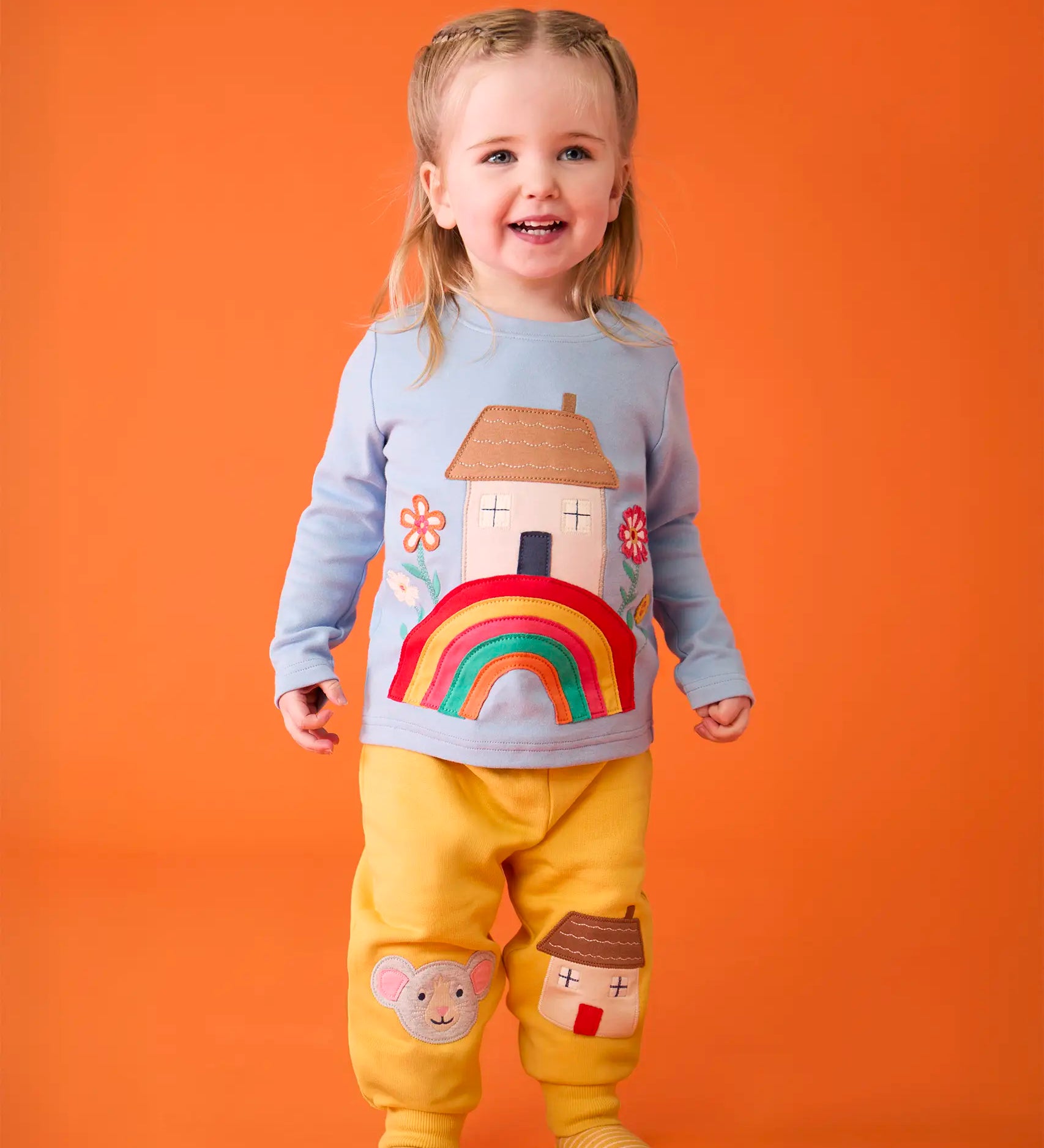 Child wearing the Frugi's GOTS 100% organic cotton top in light blue showing the cute house on a rainbow applique on the front with matching yellow character applique joggers. 