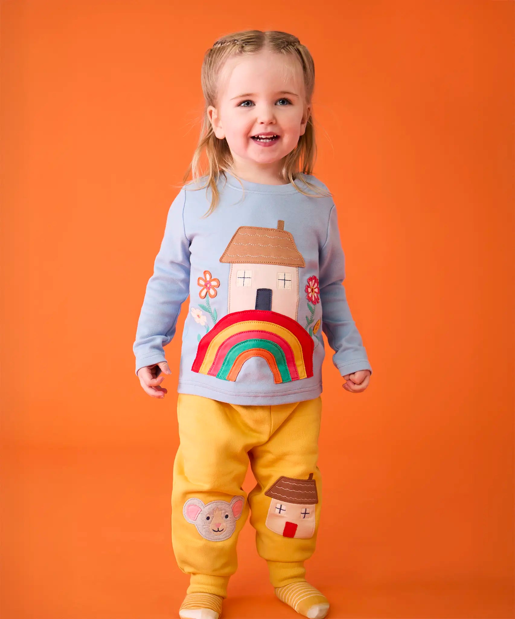 Child wearing the Frugi's GOTS 100% organic cotton top in light blue showing the cute house on a rainbow applique on the front with matching yellow character applique joggers. 