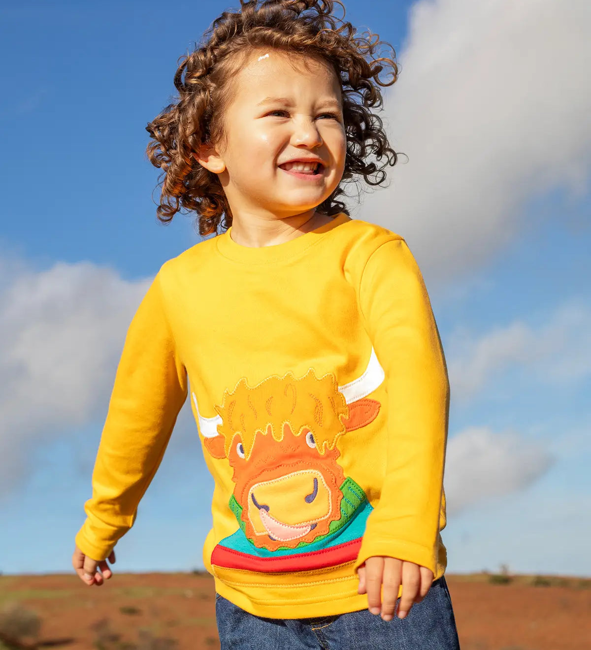 Child wearing the Frugi adventure circular cotton top in bumblebee yellow with a Highland cow applique on the front and denim jeans.