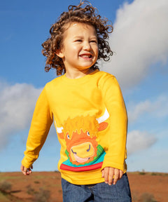 Child wearing the Frugi adventure circular cotton top in bumblebee yellow with a Highland cow applique on the front and denim jeans.