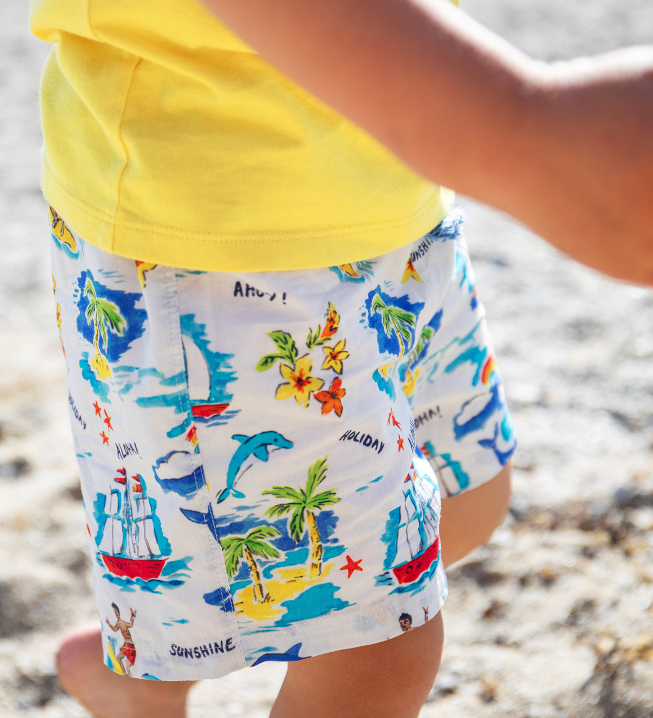 A close up of a toddler wearing the Frugi Aloha Edie Shorts featuring an all-over tropical print with sailing ship, dolphins, rainbows, palm trees, surfers and beach motifs, while running on sand.