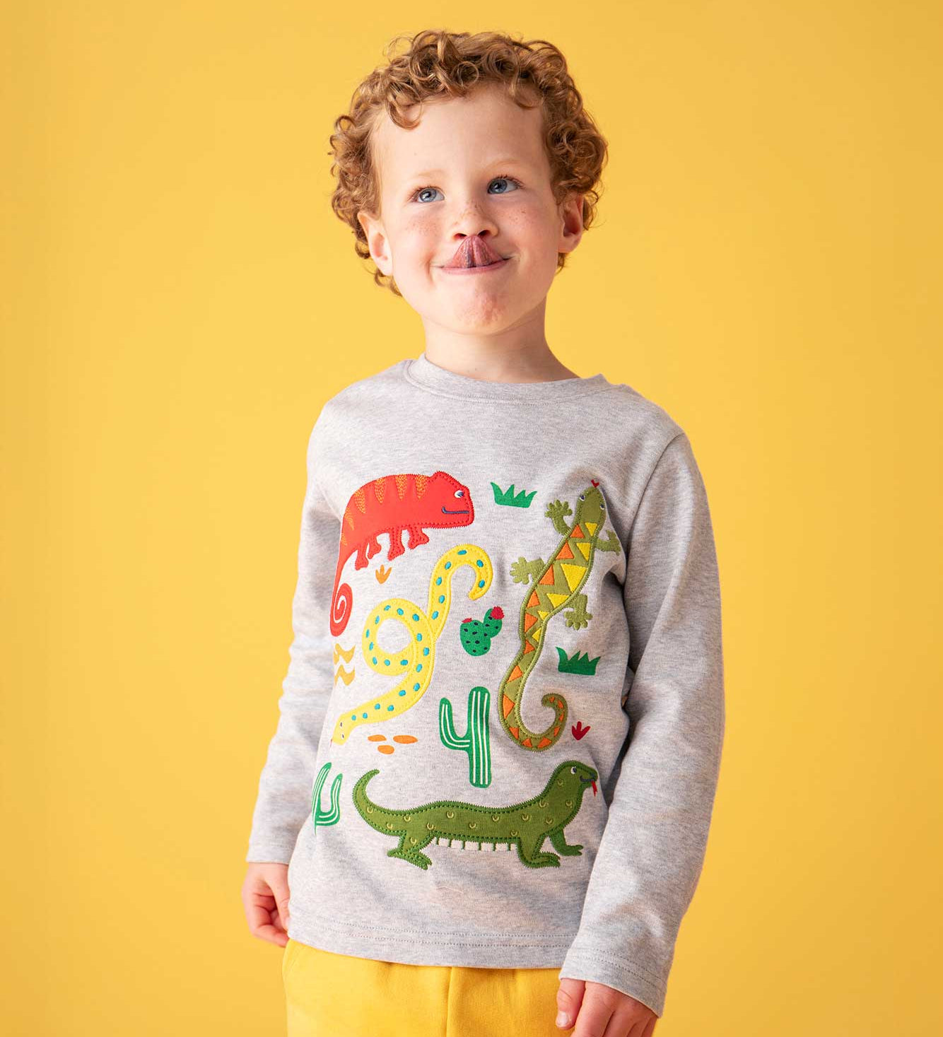 A child with blonde curly hair sticking his tounge out wearing a Frugi grey marl amphibian applique long sleeved organic cotton top. A mix of appliques and printed details featuring 4 amphibians, cacti and greenery. 