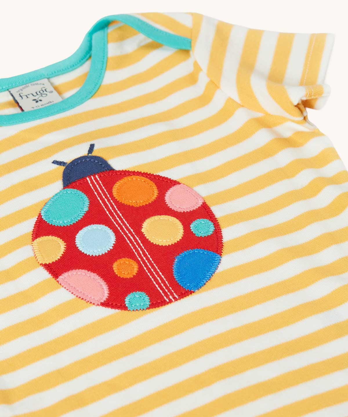 Close up of the front of the Frugi yellow striped short sleeve t-shirt with ladybird applique on the front. The ladybird has rainbow coloured dots.