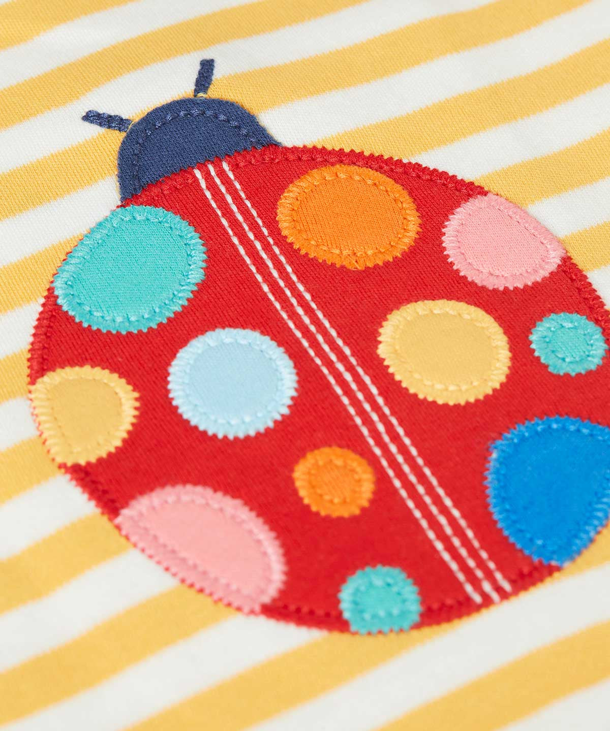 Close up of ladybird applique on the front of the Frugi yellow striped short sleeve t-shirt. The red ladybird has orange, pink, light yellow, blue and light blue coloured dots.