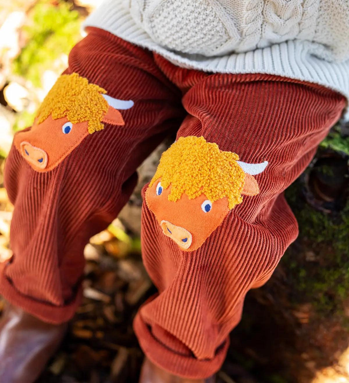 Close up of the highland cow knee patch appliques on the Frugi Arlo rust brown cord trousers, Child wearing with cable knit cream jumper.