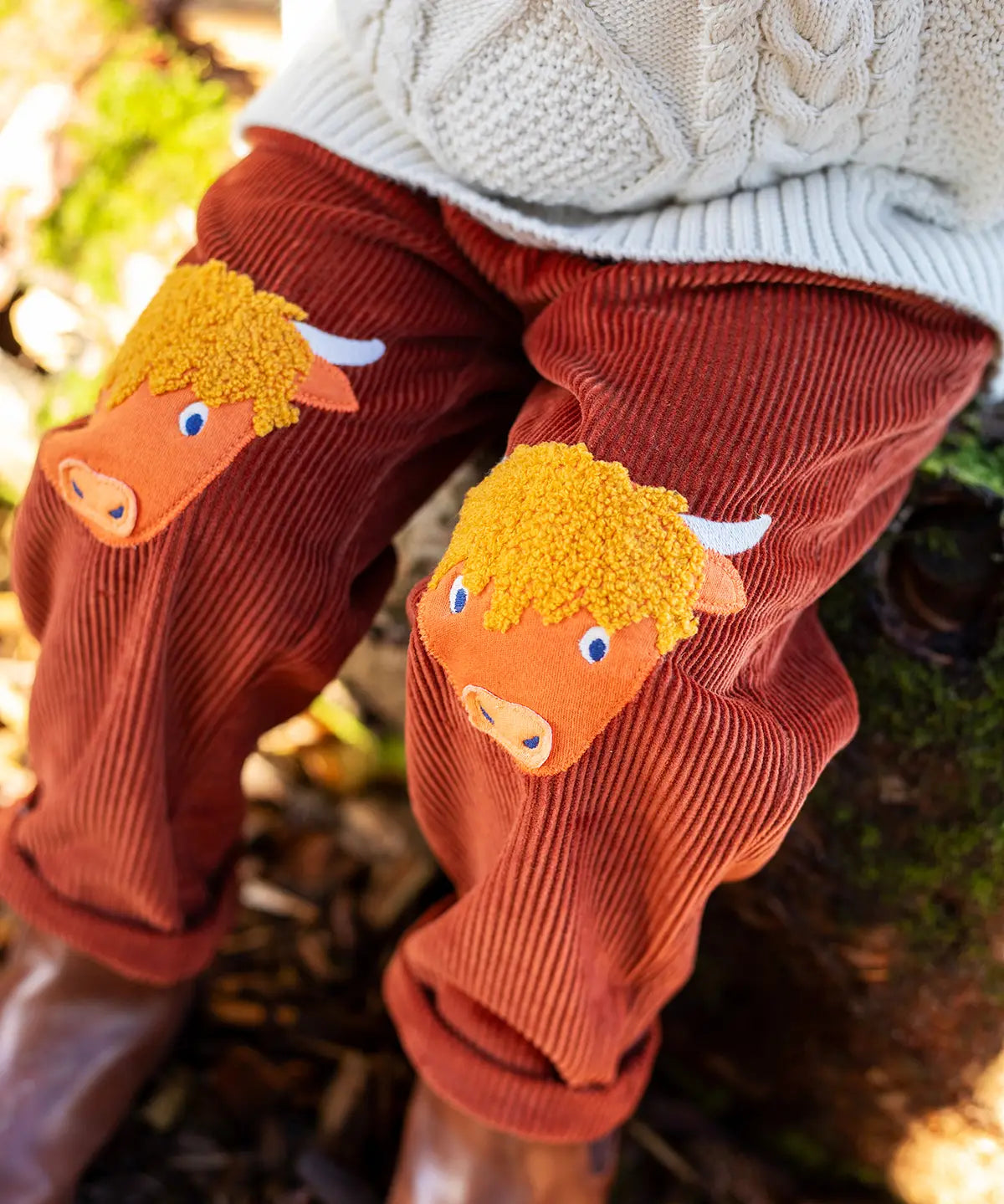 Close up of the highland cow knee patch appliques on the Frugi Arlo rust brown cord trousers, Child wearing with cable knit cream jumper.