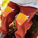 Close up of the highland cow knee patch appliques on the Frugi Arlo rust brown cord trousers, Child wearing with cable knit cream jumper.