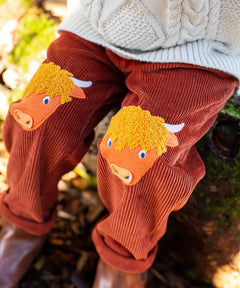 Close up of the highland cow knee patch appliques on the Frugi Arlo rust brown cord trousers, Child wearing with cable knit cream jumper.