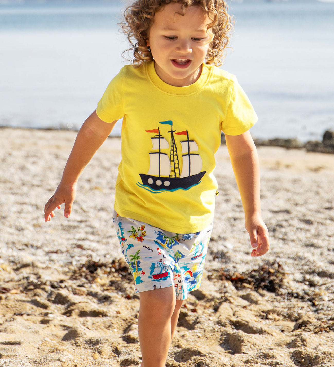 A toddler wearing Frugi Avery Appligue T-Shirt in Welly Yellow, with a Tall Ship embroidered design in the centre, running on a sandy beach.