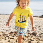 A toddler wearing Frugi Avery Appligue T-Shirt in Welly Yellow, with a Tall Ship embroidered design in the centre, running on a sandy beach.