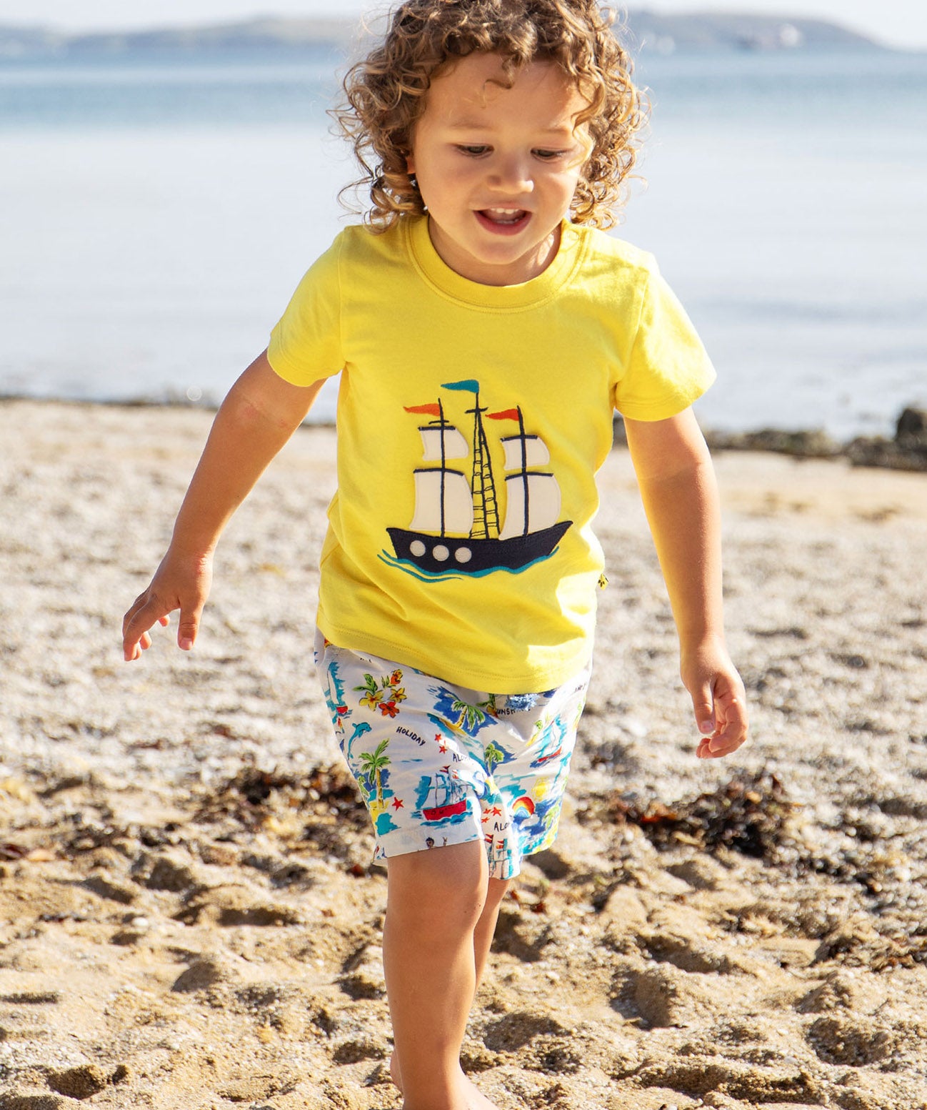 A toddler wearing Frugi Avery Appligue T-Shirt in Welly Yellow, with a Tall Ship embroidered design in the centre, running on a sandy beach.