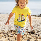 A toddler wearing Frugi Avery Appligue T-Shirt in Welly Yellow, with a Tall Ship embroidered design in the centre, running on a sandy beach.