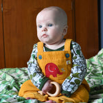 A baby wearing the Frugi Babipur yellow cord dungarees sitting on a bed with a wooden toy teether in his hand. 