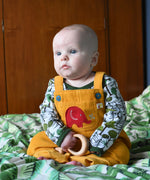 A baby wearing the Frugi Babipur yellow cord dungarees sitting on a bed with a wooden toy teether in his hand. 