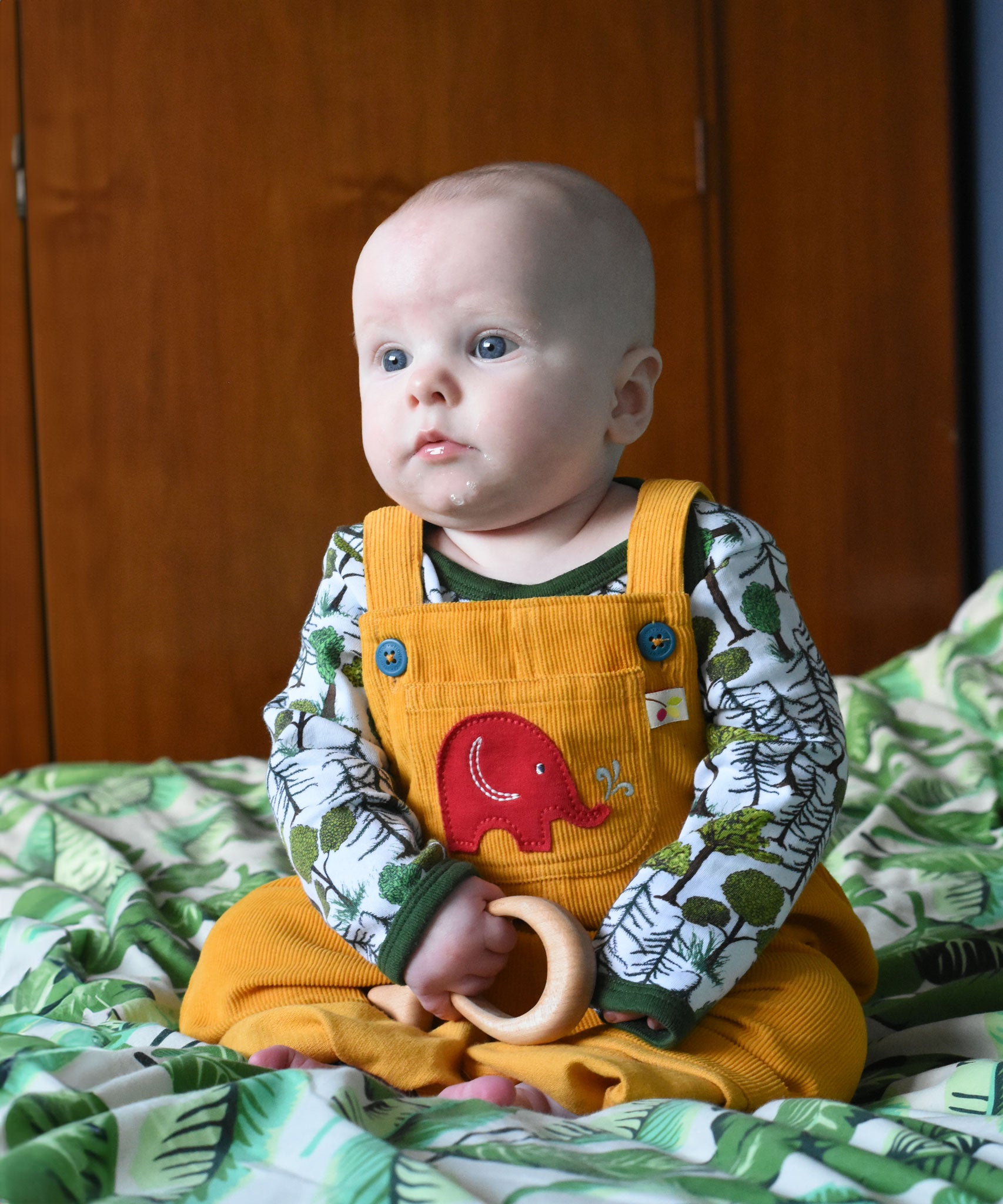A baby wearing the Frugi Babipur yellow cord dungarees sitting on a bed with a wooden toy teether in his hand. 