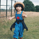 A child wearing teh blue Frugi Babipur cord dungarees with a straw hat and a black checked shirt tied around his waist. 